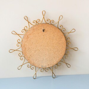 Puede incluir: Un espejo vintage con forma de sol con un marco de metal dorado. La parte posterior del espejo es un panel redondo de madera marr&oacute;n claro. El marco presenta un dise&ntilde;o intrincado y arremolinado, creando una apariencia de sol. Un peque&ntilde;o gancho de metal est&aacute; unido a la parte posterior para colgar.