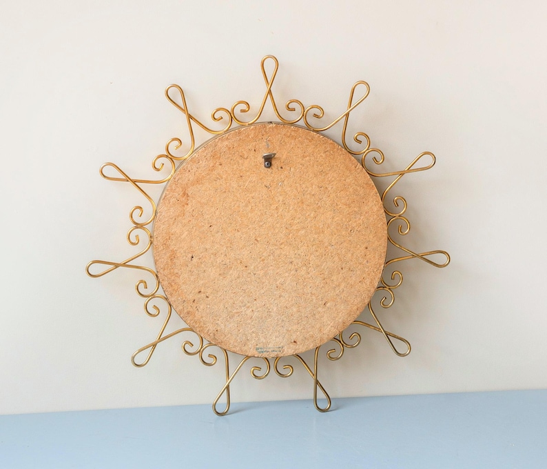 Puede incluir: Un espejo vintage con forma de sol con un marco de metal dorado. La parte posterior del espejo es un panel redondo de madera marr&oacute;n claro. El marco presenta un dise&ntilde;o intrincado y arremolinado, creando una apariencia de sol. Un peque&ntilde;o gancho de metal est&aacute; unido a la parte posterior para colgar.