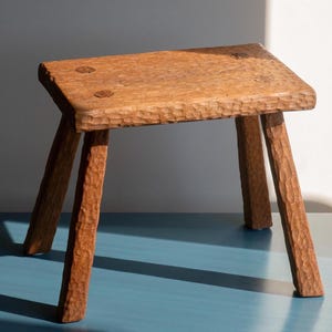 Può includere: Uno sgabello in legno rustico con un piano grezzo e tre gambe. Lo sgabello è realizzato in legno naturale e ha un colore marrone scuro.