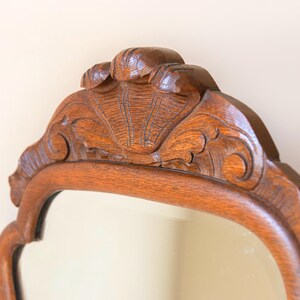 May include: A wall mirror featuring a medium-brown wooden frame with a polished finish. The top section of the frame displays an intricate, carved shell motif centred within a decorative, curved wooden crest that extends across the upper edge.