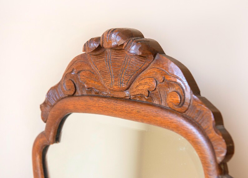 May include: A wall mirror featuring a medium-brown wooden frame with a polished finish. The top section of the frame displays an intricate, carved shell motif centred within a decorative, curved wooden crest that extends across the upper edge.
