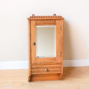 May include: A vintage wooden medicine cabinet with a mirrored door and a small drawer. The cabinet is made of light brown wood and features a decorative top edge and brass hardware. It is mounted on a wall.