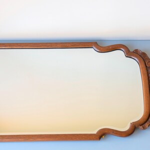 May include: A rectangular wall mirror with a medium brown wooden frame. The frame features a smooth straight edge on three sides and a decorative carved wooden crest with shell-like detailing on the right side, creating a unique silhouette.
