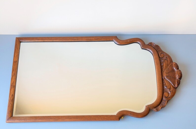 May include: A rectangular wall mirror with a medium brown wooden frame. The frame features a smooth straight edge on three sides and a decorative carved wooden crest with shell-like detailing on the right side, creating a unique silhouette.