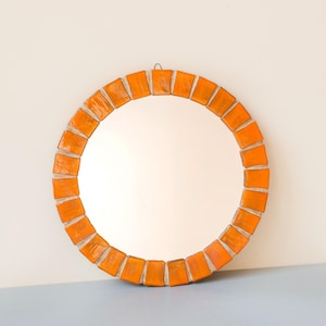 May include: A circular wall mirror featuring a decorative frame composed of individual, glossy orange rectangular tiles arranged in a radial pattern. The mirror has a smooth, reflective glass centre and a small metal hanging loop at the top.