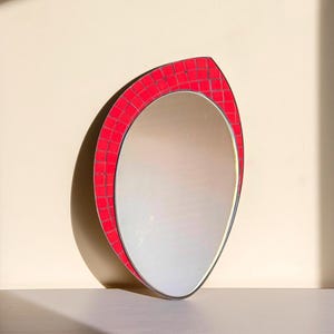 May include: A teardrop-shaped wall mirror featuring a vibrant red mosaic tile border. The reflective glass surface is framed by small, square, bright red tiles arranged in a grid pattern, creating a distinct, pointed, asymmetrical silhouette.