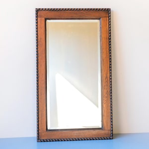 Op de afbeelding: Rechthoekige spiegel met een houten frame en een touwachtige rand. De lijst is bruin en het glas reflecteert het licht. De spiegel is een verticaal rechthoek, geschikt voor slaapkamer of badkamer.