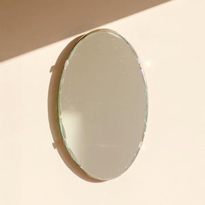 Peut inclure: Miroir ovale avec un bord biseauté, monté sur un mur de couleur claire. Le miroir reflète l'environnement, et est maintenu en place par de petits supports discrets. Le design est simple et minimaliste, adapté à divers styles d'intérieur.