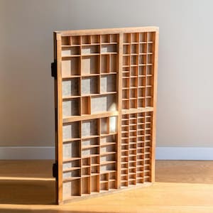May include: A vintage wooden printer's tray with numerous small compartments. The tray is made of light-colored wood and features a grid-like design. It is likely used for organizing small items or printing blocks. The tray is leaning against a wall.