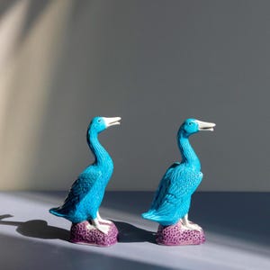 Op de afbeelding: Twee turquoise keramische vogelbeeldjes, elk staand op een paarse, getextureerde basis. De vogels hebben witte snavels en zijn symmetrisch geplaatst. Deze decoratieve beeldjes voegen een vleugje kleur toe aan elke ruimte. Hoogte ongeveer 15 cm.