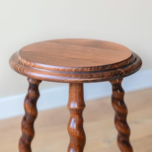 May include: A round wooden stool with a smooth, flat top and three distinctively carved, spiraling legs. The wood features a rich, medium-brown finish with visible grain patterns throughout the surface of the circular seat and the twisted legs.