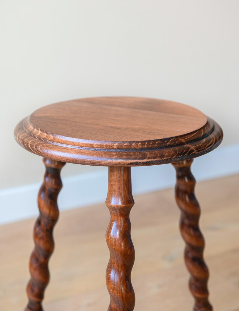 May include: A round wooden stool with a smooth, flat top and three distinctively carved, spiraling legs. The wood features a rich, medium-brown finish with visible grain patterns throughout the surface of the circular seat and the twisted legs.