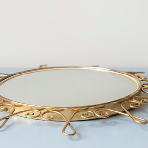 Puede incluir: Un espejo de tocador redondo vintage con un marco de metal dorado. El marco presenta un dise&ntilde;o ornamental de rayos de sol con remolinos decorativos y detalles en forma de l&aacute;grima. Es un art&iacute;culo decorativo.