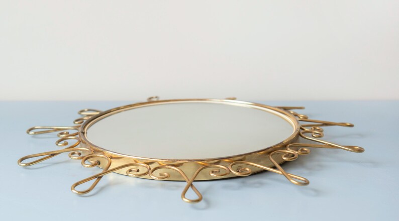 Puede incluir: Un espejo de tocador redondo vintage con un marco de metal dorado. El marco presenta un dise&ntilde;o ornamental de rayos de sol con remolinos decorativos y detalles en forma de l&aacute;grima. Es un art&iacute;culo decorativo.