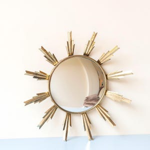 May include: A circular sunburst mirror with a gold-coloured frame. The mirror has a reflective surface surrounded by pointed rays. The frame and rays have a metallic finish. The mirror is a decorative wall hanging.