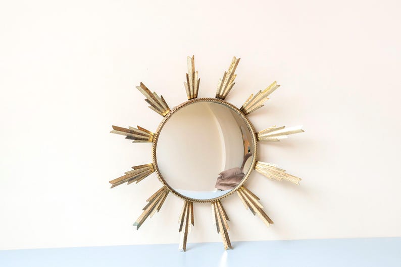 May include: A circular sunburst mirror with a gold-coloured frame. The mirror has a reflective surface surrounded by pointed rays. The frame and rays have a metallic finish. The mirror is a decorative wall hanging.