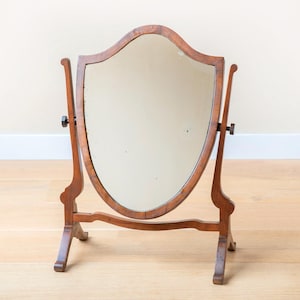 Puede incluir: Un espejo de tocador antiguo de madera, con forma de escudo. El espejo está enmarcado en madera marrón oscuro y está sostenido por dos brazos de madera curvados y una base. El espejo es ajustable y se apoya sobre cuatro patas. La madera tiene una veta natural.