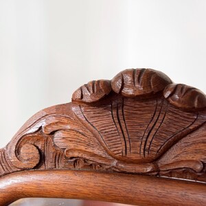May include: A dark brown wooden furniture crest featuring intricate hand-carved details. The top centre displays a prominent shell motif flanked by smaller carved elements, with scrolling patterns extending along the curved frame edge.