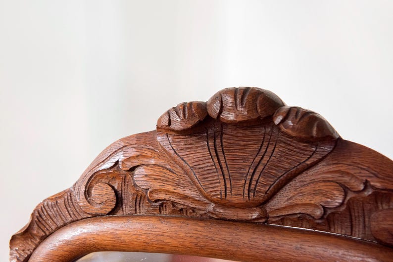 May include: A dark brown wooden furniture crest featuring intricate hand-carved details. The top centre displays a prominent shell motif flanked by smaller carved elements, with scrolling patterns extending along the curved frame edge.