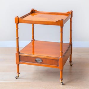 Könnte beinhalten: Ein zweistöckiger Beistelltisch aus Holz mit warmem, rotbraunem Finish. Der Tisch hat eine Schublade mit Metallgriff, Beine im Bambus-Stil und kleine Rollen. Der Tisch ist etwa 71 cm hoch.