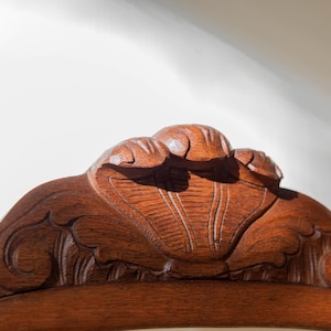 May include: A close-up of the top crest of a wooden chair featuring intricate hand-carved details. The reddish-brown wood displays a visible grain pattern with a central shell-like motif flanked by symmetrical scrolling carvings along the edges.