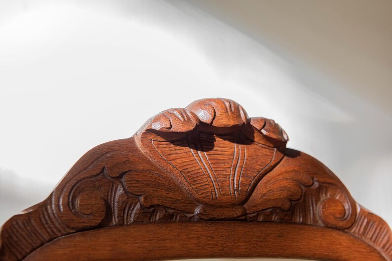 May include: A close-up of the top crest of a wooden chair featuring intricate hand-carved details. The reddish-brown wood displays a visible grain pattern with a central shell-like motif flanked by symmetrical scrolling carvings along the edges.