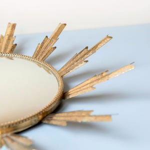May include: A gold sunburst mirror with a circular reflective centre. The frame has radiating, pointed rays. The design is reminiscent of Art Deco style. The mirror is set against a light blue background.