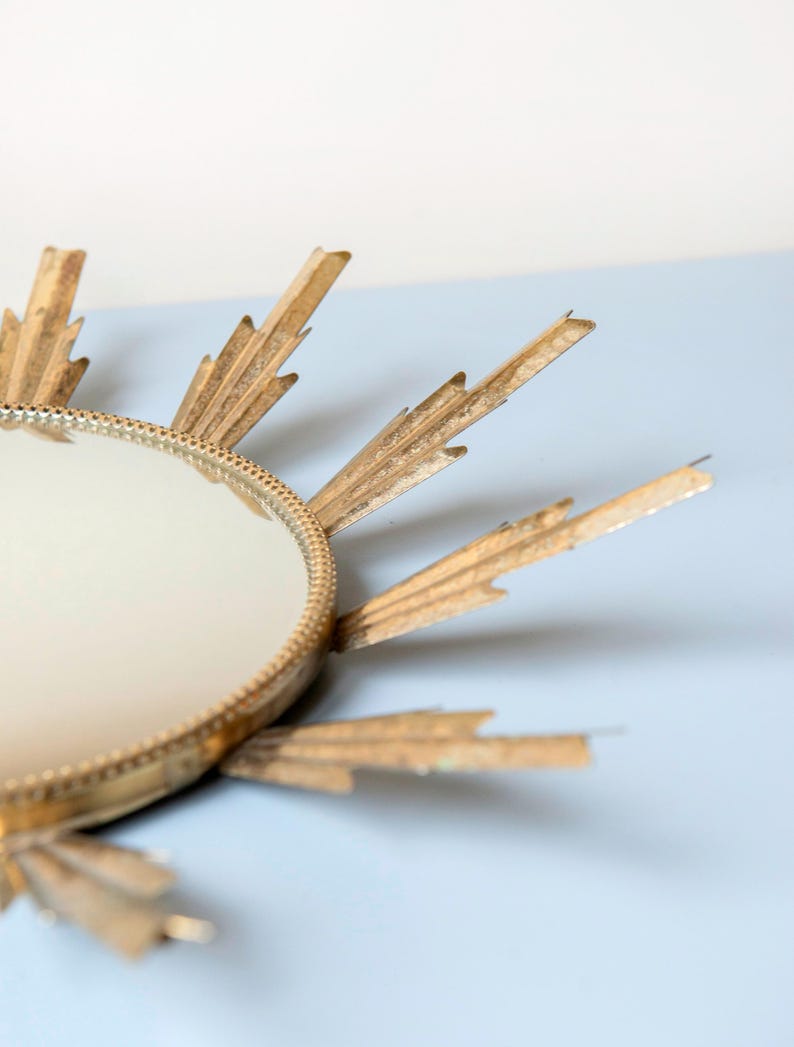 May include: A gold sunburst mirror with a circular reflective centre. The frame has radiating, pointed rays. The design is reminiscent of Art Deco style. The mirror is set against a light blue background.
