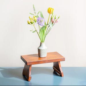 Puede incluir: Un pequeño taburete de madera con un acabado marrón claro y un jarrón con un ramo de flores amarillas, rosas y moradas. El jarrón es de color gris claro y tiene una superficie texturizada.