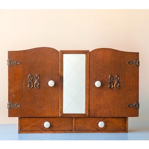 Op de afbeelding: Een houten wandkast met twee deuren voorzien van sierlijke metalen scharnieren en decoratieve metalen platen, een centrale verticale spiegel en twee onderste lades. Elke deur en lade is uitgerust met een kleine, ronde, witte keramische knop.