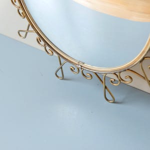 Puede incluir: Espejo ovalado con marco de metal dorado. El marco presenta adornos decorativos y est&aacute; sostenido por delicadas patas curvas. El espejo est&aacute; sobre una superficie azul claro.