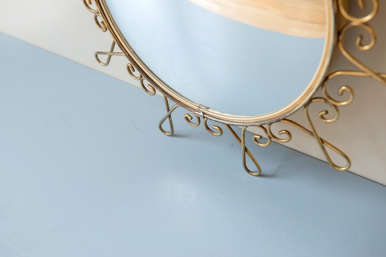 Puede incluir: Espejo ovalado con marco de metal dorado. El marco presenta adornos decorativos y est&aacute; sostenido por delicadas patas curvas. El espejo est&aacute; sobre una superficie azul claro.