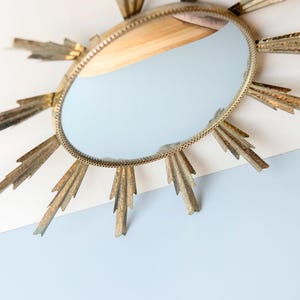 May include: Oval gold sunburst mirror with a light blue reflection. The mirror has a gold frame with a beaded edge and gold rays extending outwards. The mirror is mounted on a white and light blue surface.