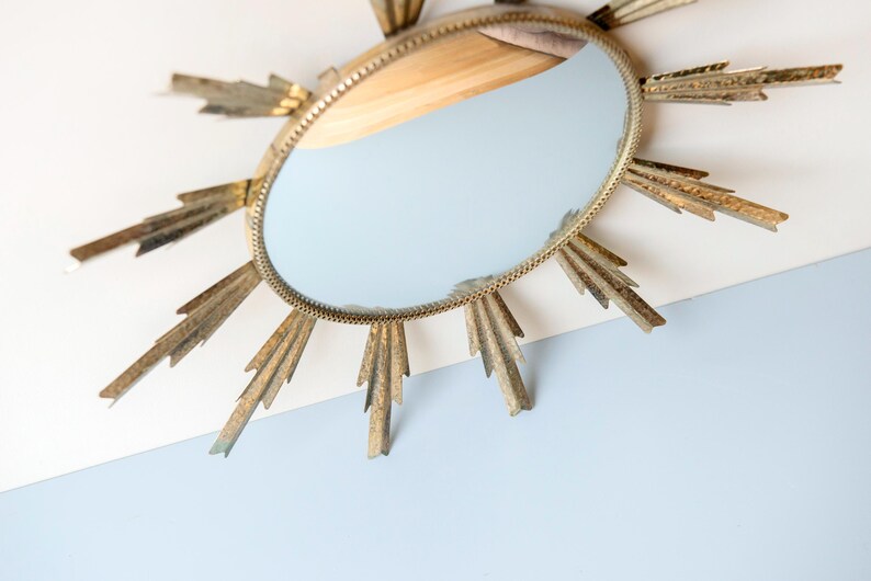 May include: Oval gold sunburst mirror with a light blue reflection. The mirror has a gold frame with a beaded edge and gold rays extending outwards. The mirror is mounted on a white and light blue surface.