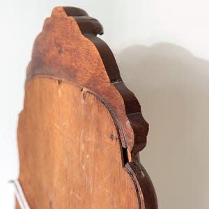 May include: A wooden mirror frame with a scalloped, curved top edge and a dark brown finish. The back panel is made of a lighter, smooth, unfinished wood material. The frame features a distinct layered construction with visible wood grain.
