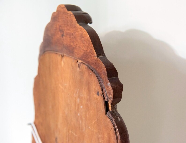 May include: A wooden mirror frame with a scalloped, curved top edge and a dark brown finish. The back panel is made of a lighter, smooth, unfinished wood material. The frame features a distinct layered construction with visible wood grain.
