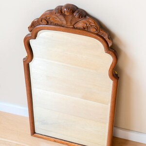 May include: A wall mirror featuring a medium-brown wooden frame with a smooth finish. The top of the frame is arched and decorated with carved, fan-like detailing. The mirror has a rectangular glass pane with curved upper corners and straight sides.