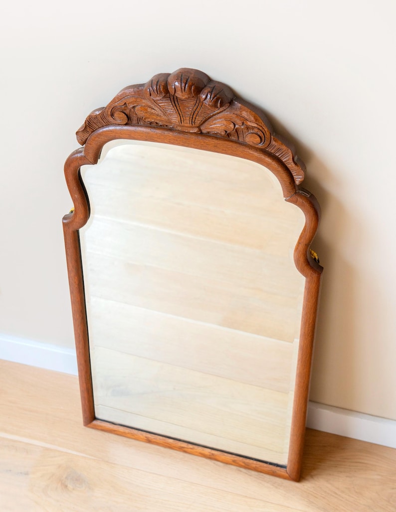 May include: A wall mirror featuring a medium-brown wooden frame with a smooth finish. The top of the frame is arched and decorated with carved, fan-like detailing. The mirror has a rectangular glass pane with curved upper corners and straight sides.