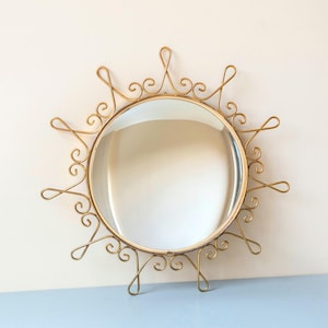 Puede incluir: Espejo redondo de metal dorado con un marco ornamental en forma de sol. El marco presenta dise&ntilde;os en espiral y bucles delicados. El espejo es un elemento decorativo para colgar en la pared.