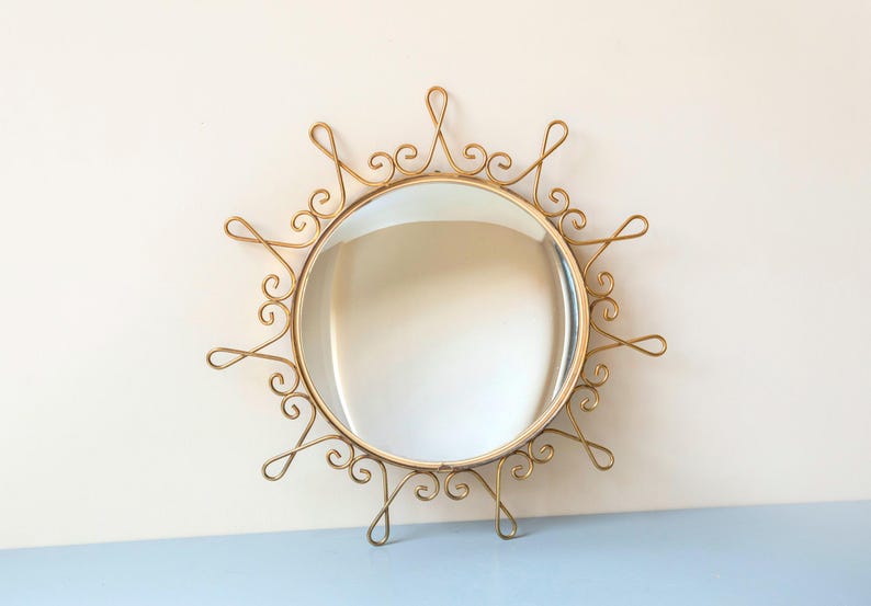 Puede incluir: Espejo redondo de metal dorado con un marco ornamental en forma de sol. El marco presenta dise&ntilde;os en espiral y bucles delicados. El espejo es un elemento decorativo para colgar en la pared.