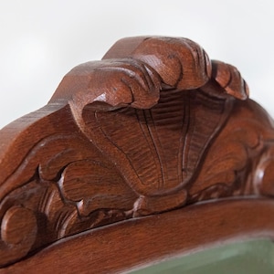 May include: A dark brown wooden furniture crest featuring a hand-carved shell motif at the centre, flanked by symmetrical scrolling leaf patterns. The wood grain is visible across the polished surface, showcasing detailed relief carvings.