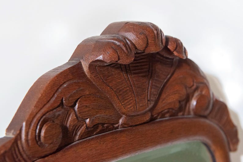 May include: A dark brown wooden furniture crest featuring a hand-carved shell motif at the centre, flanked by symmetrical scrolling leaf patterns. The wood grain is visible across the polished surface, showcasing detailed relief carvings.
