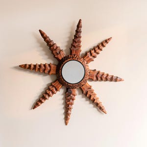 Puede incluir: Espejo de madera con forma de sol, con un espejo circular central y puntas texturizadas que irradian. El espejo es de madera marrón cálido y está montado en una pared de color neutro. El diseño evoca el sol o una estrella.