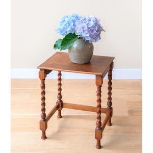 Peut inclure: Petite table d'appoint carrée en bois avec des pieds torsadés sculptés. Un vase en céramique grise contient un bouquet d'hortensias bleus et violets. La table mesure environ 61 cm de haut.