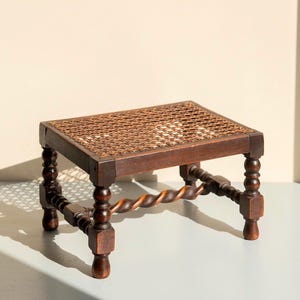 Op de afbeelding: Een kleine, antieke houten voetenbank met een geweven rieten bovenkant. Het donkerbruine frame heeft gedraaide poten en een decoratieve, gedraaide ligger. De voetenbank is een klassiek meubelstuk.