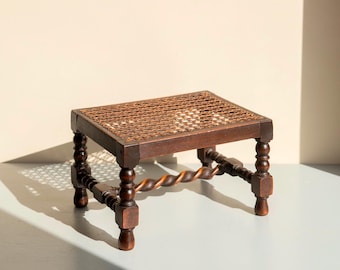 May include: A small, antique wooden footstool with a woven cane top. The dark brown frame features turned legs and a decorative twisted stretcher. The footstool is a classic piece of furniture.