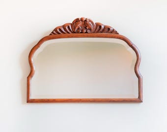 Puede incluir: Espejo decorativo de estilo antiguo con marco de madera tallada. El marco es de color marrón cálido y presenta un diseño ornamental tallado en la parte superior. El espejo tiene forma curva y está montado en una pared blanca.