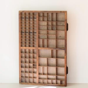 May include: A rectangular wooden printer's tray featuring a grid of numerous small, open compartments of varying sizes. The wood has a natural brown finish with visible grain and signs of wear, and the structure is divided into four main sections.