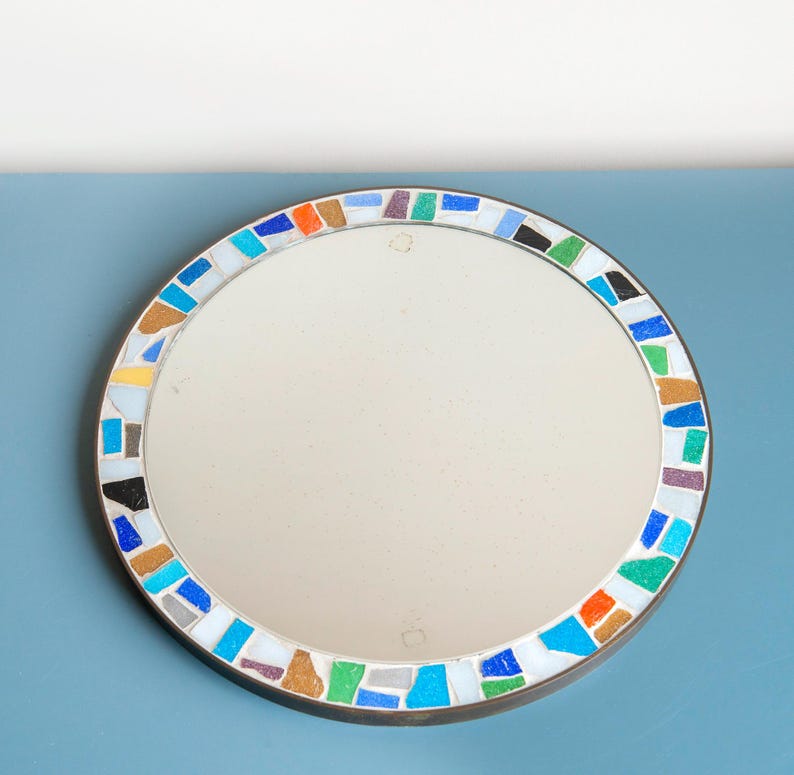May include: A round mirror with a colorful mosaic border. The mosaic is made of small, irregular pieces of glass in shades of blue, green, yellow, orange, and brown. The mirror is framed in a thin, brass-colored metal.