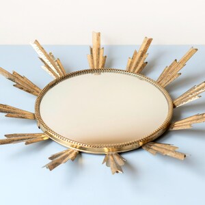 May include: A gold-coloured sunburst mirror with a round, reflective centre. The mirror's frame features radiating, stylised rays. The design is vintage-inspired, adding a decorative element to any space. The mirror is a decorative wall hanging.